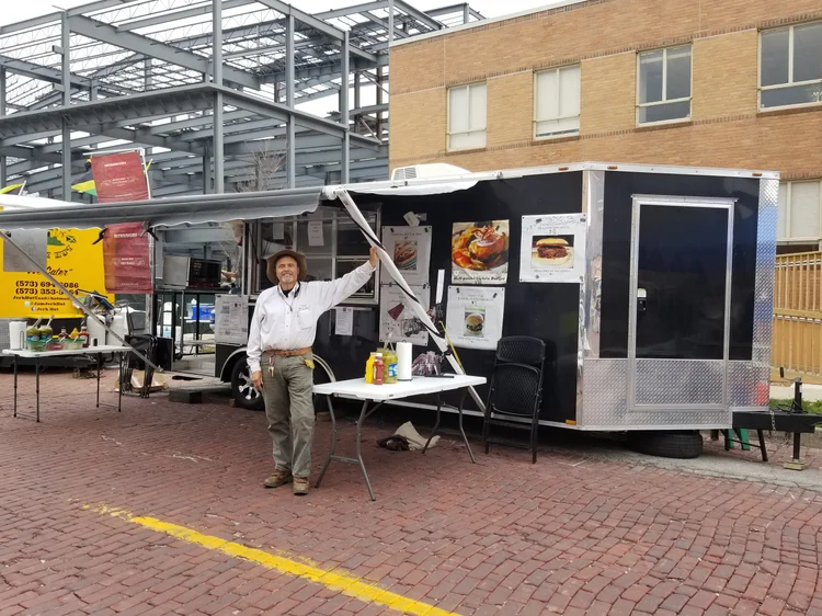 20180422_111432 Food truck owner standing outside a mobile food trailer, showcasing menu items and condiments on a table, with a construction site in the background.