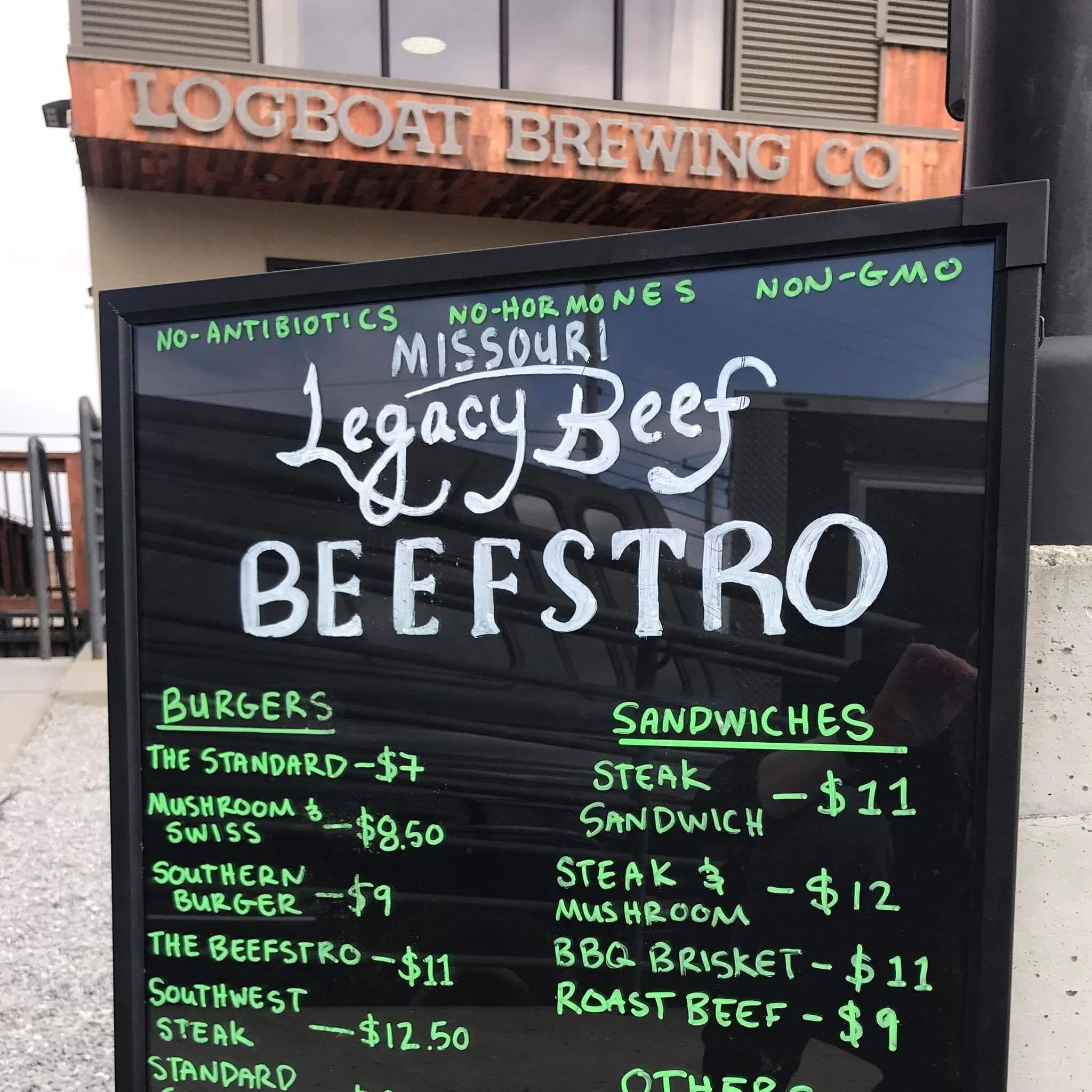FB_IMG_1557097561508 Menu board outside Logboat Brewing Co. featuring Missouri Legacy Beef Beefstro offerings, including burgers and sandwiches. Highlights include The Standard burger for $7, Mushroom & Swiss burger for $8.50, and a selection of sandwiches like Steak Sandwich and BBQ Brisket, all emphasizing no antibiotics, no hormones, and non-GMO ingredients.
