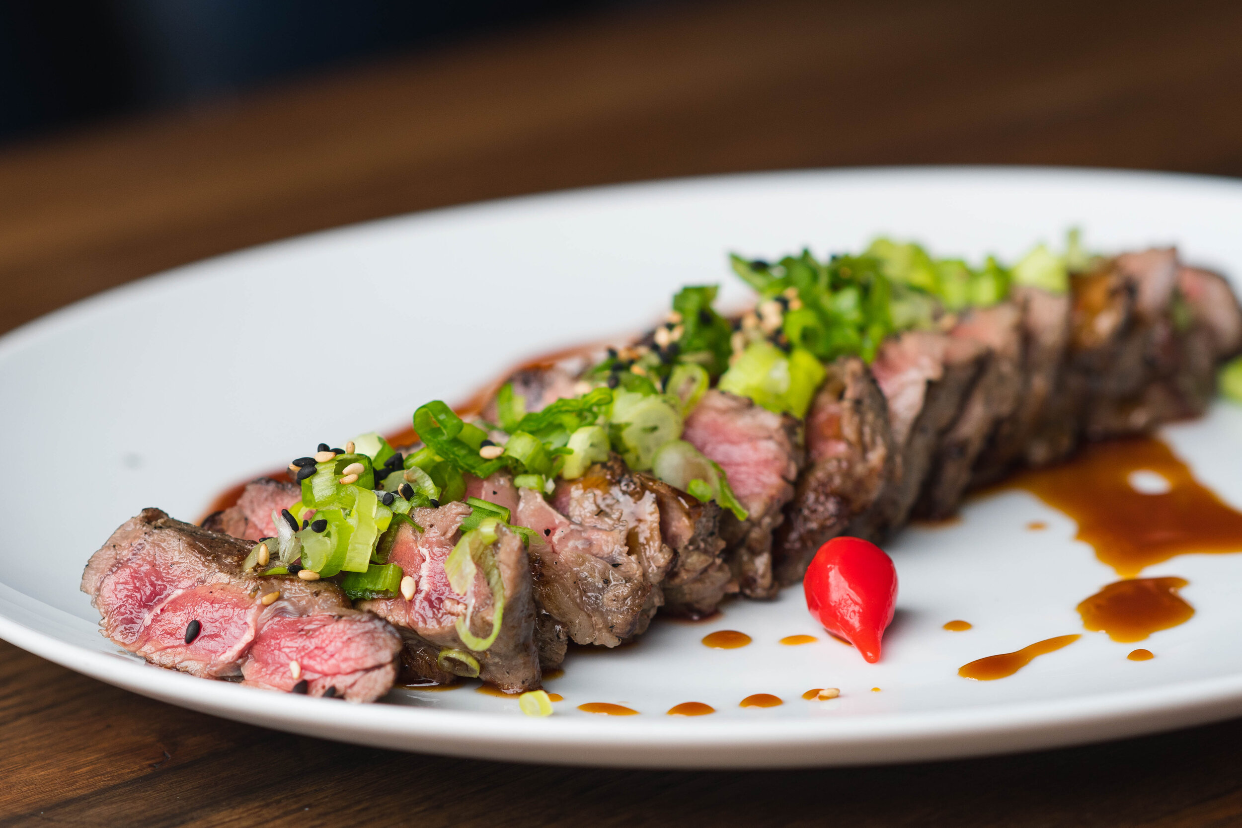 image-asset (1) Sliced steak garnished with green onions and sesame seeds, drizzled with sauce, served on a white plate. A bright red chili pepper adds a pop of color to the dish.