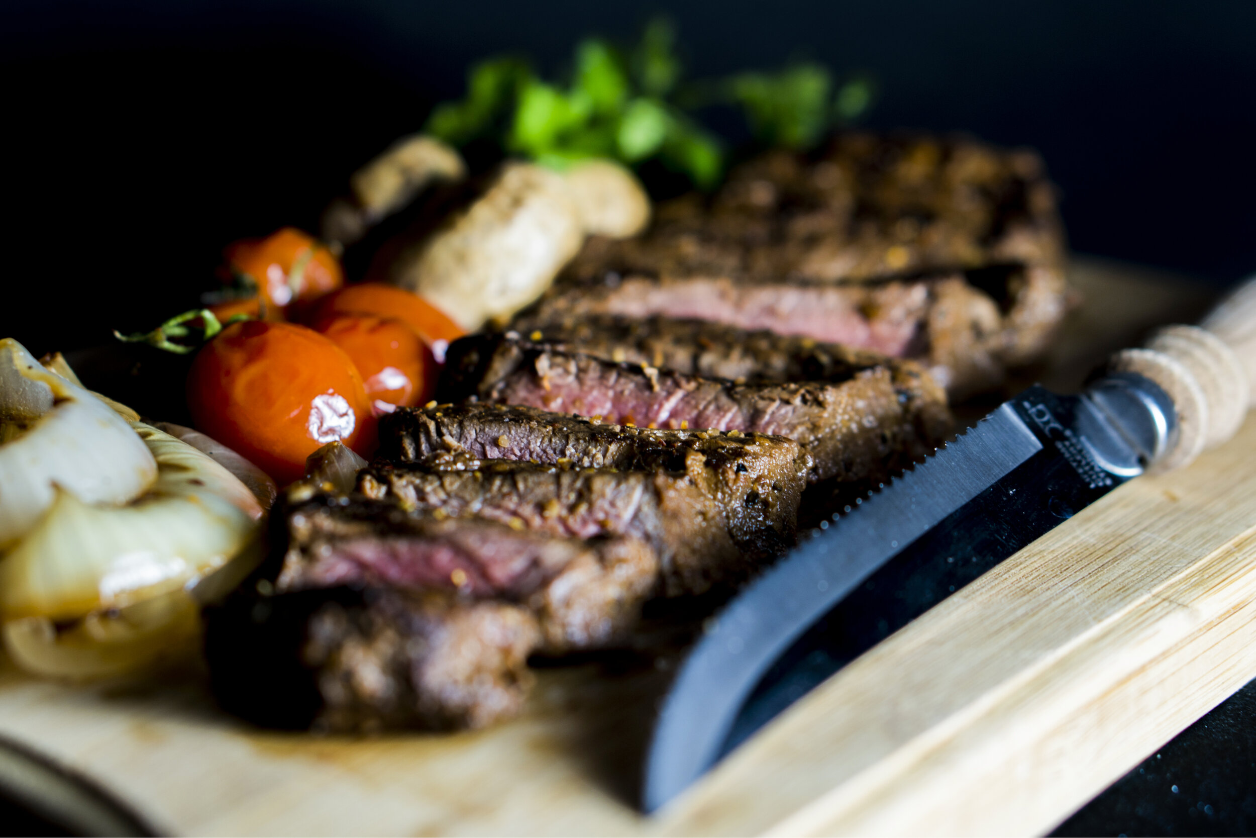 Juicy grilled steak slices on a wooden cutting board, accompanied by roasted cherry tomatoes, sautéed onions, and mushrooms, with a serrated knife for serving.