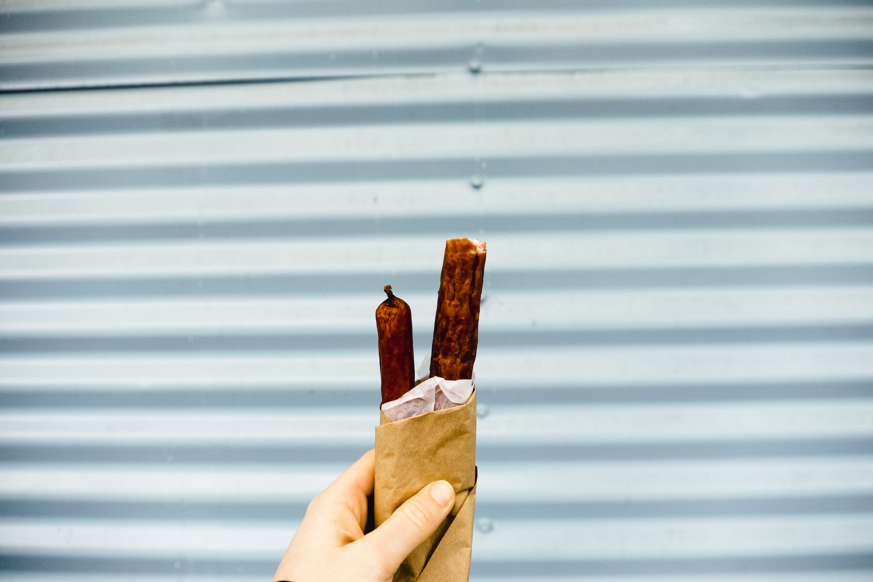 image-asset (3) Hand holding two pieces of sausage wrapped in paper against a blue corrugated metal background.