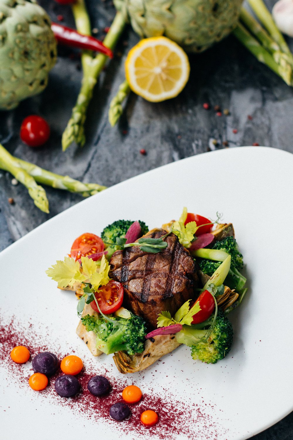image-asset (4) Grilled steak served on a bed of sautéed broccoli, cherry tomatoes, and artichokes, garnished with fresh herbs and colorful vegetable purees on a white plate, surrounded by fresh asparagus and lemon.